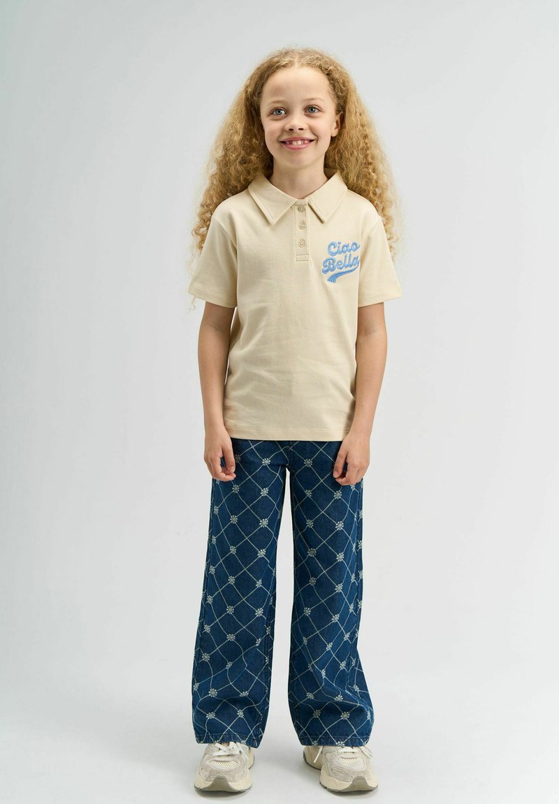 Polo beige avec "Ciao Bella" brodé en bleu, assorti à un pantalon en denim foncé à jambe large, présentant un motif en losange et de légacs accents floraux.