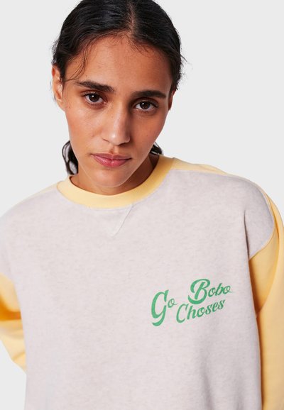 Woman with dark hair wearing a light gray and yellow sweatshirt with green "Go Bobo Chooses" text, looking directly at the camera.