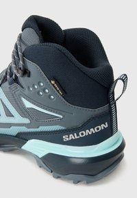 Pusē augstas pārgājienu zābaki zilos un pelēkos toņos. Iekļauj mežģīņu paneļus, strukturētus apvalkus, izteiktu logotipu un GORE-TEX zīmolu.
