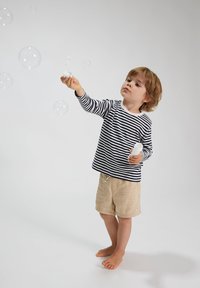 Gestreiftes Langarmhemd mit Marine- und weißen Streifen, beige Shorts, Kind bläst Seifenblasen. Barfuß, heller Hintergrund, verspielte Szene.
