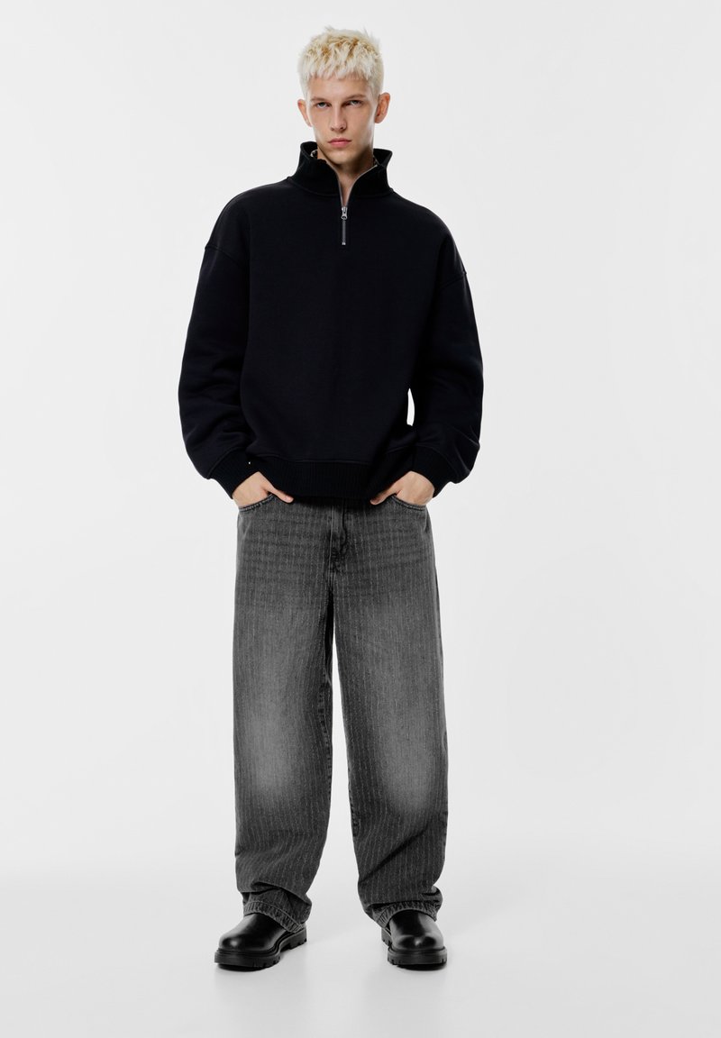 Felpa nera con zip e collo alto, abbinata a jeans grigi a righe a gamba larga e scarpe nere, in piedi davanti a uno sfondo bianco.