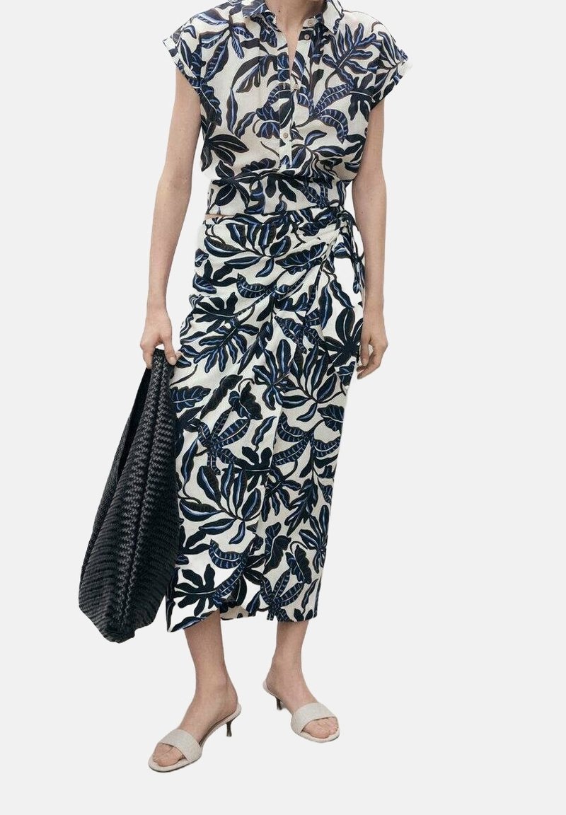 Femme portant une robe blanche avec des motifs de feuilles bleues et noires, tenant un grand sac noir tressé, et portant des sandales blanches à petit talon.