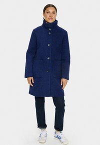 Marinblå ullkappa med hög krage, framknäppning och två stora fickor. Strukturen på tyget är lös och passar bra tillsammans med mörka jeans och sneakers.