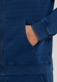 Felpa con zip blu navy e finitura texturizzata, dotata di tasca frontale e polsini a costine. Tessuto liscio e dettagli di cucitura minimi.