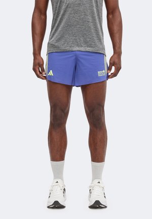 Homme portant un t-shirt gris, un short bleu BMW Berlin Marathon, des chaussettes blanches et des chaussures de course blanches Adidas, debout devant un fond uni.