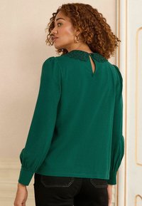 Grüne Bluse mit strukturiertem Spitzenkragen und Schlüssellochausschnitt. Mit Puffärmeln und glattem Stoff, kombiniert mit schwarzen Hosen.