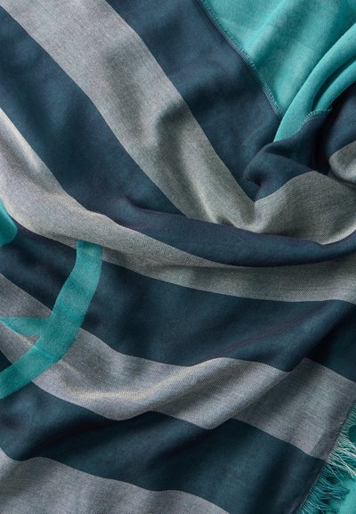 Απαλή υφασμάτινη ύλη με φαρδιές οριζόντιες ρίγες σε αποχρώσεις του μπλε και του γκρι, διπλωμένη με ορατή υφή και κρόσσια στην άκρη πάνω σε τυρκουάζ φόντο.