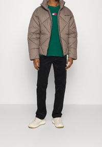 Brun quiltad pufferjacka med hög krage, bärs över en grön T-shirt med logotyp, kombinerad med svarta byxor och vita sneakers.