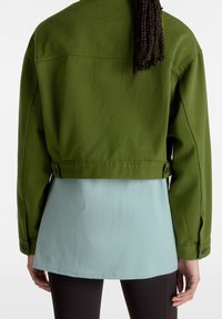 Giacca verde oliva tagliata con una texture liscia, caratterizzata da dettagli con bottoni e una maglietta blu, posizionata su uno sfondo bianco uniforme.