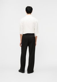 Άνδρας που στέκεται με την πλάτη γυρισμένη, φοράει λευκό πουκάμισο με μακριά μανίκια και γυρισμένα μανικέτια, μαύρο παντελόνι και μαύρα παπούτσια σε λευκό φόντο.