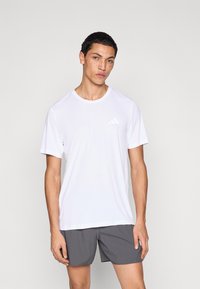 Camiseta deportiva blanca de manga corta en tejido ligero, con cuello redondo y logo sutil en el pecho, combinada con pantalones cortos grises.
