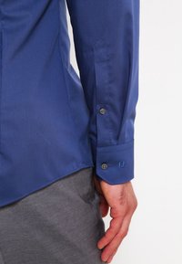 Blauwe, langmouwige shirt gemaakt van gladde stof. Heeft een puntige manchet met twee knopen en een subtiele textuur. Gemaakt om te dragen met grijze broek.