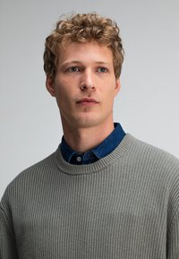 Grijze geribbelde trui met een ronde hals die over een blauwe denim overhemd is gedragen. Heeft een gestructureerde stof en een relaxte pasvorm.