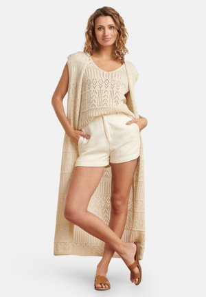 Beige knitted vest with open front, matching textured tank top, cream shorts, and brown slide sandals. Patterns visible on vest design.
