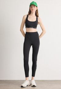Melnas sporta krūšturis un augsta jostasvietas legingi ar baltu "Calvin Klein" logo. Modelis valkā zaļu cepuri un baltas sporta kurpes. Gluda auduma tekstūra.