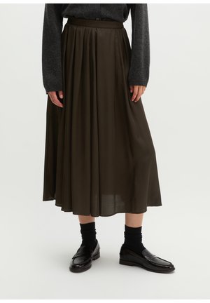 Frau trägt einen dunkelolivgrünen plissierten Midirock, schwarze Socken, glänzende schwarze Loafers und ein dunkelgraues langärmliges Oberteil auf weißem Hintergrund.