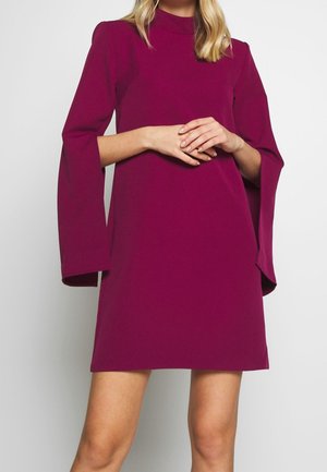 Femme portant une robe courte à manches longues de couleur magenta foncé avec de larges fentes aux manches, debout devant un fond clair uni.