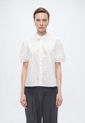 Kobieta ubrana w białą bluzkę na guziki z krótkim rękawem, ozdobioną teksturowanymi kropeczkami, oraz czarne spodnie szerokie nogawki, stojąca na jednolitym białym tle.