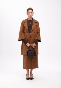 Hnědý velký zamšelý kabát s velkými knoflíky, spárovaný s přiléhavým hnědým topem a midi sukní. Drží malou černou kabelku s pleteným detailem.