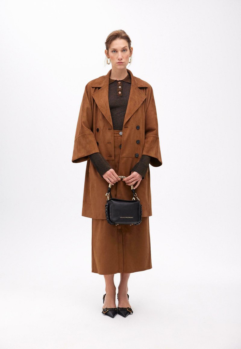 Hnědý velký zamšelý kabát s velkými knoflíky, spárovaný s přiléhavým hnědým topem a midi sukní. Drží malou černou kabelku s pleteným detailem.
