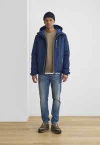 Navy blue jacket with a zip, lined hood, worn over a beige sweatshirt, paired with light blue jeans and brown hiking shoes on wooden floor.