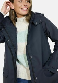 Parka imperméable bleu marine avec fermeture éclair et boutons-pression, capuche ajustable et poches latérales. Porté par-dessus un pull crème à rayures.
