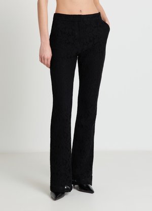 Pantalon en dentelle noire slim avec poches avant porté par une personne debout, associé à des talons pointus noirs sur un fond uni.