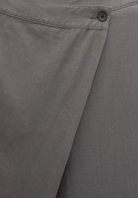 Primer plano de una tela gris con un botón negro que cierra un detalle de solapa superpuesta en la ropa.