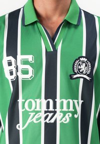 Green, black, and white vertical striped polo shirt with a collar, "85" number, tommy jeans logo text, and a crest emblem on the chest.