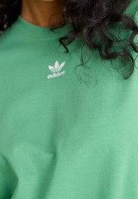 Detailní záběr na zelenou mikinu Adidas s logem na hrudi a tmavé kudrnaté vlasy přes rameno.