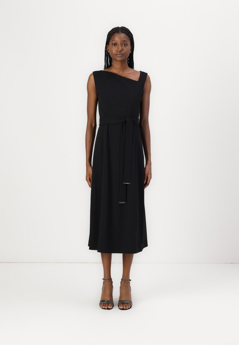 Robe noire sans manches avec un décolleté diagonal, un corsage ajusté et une ceinture à nouer. Texture lisse et longueur midi, associée à des talons à lanières.