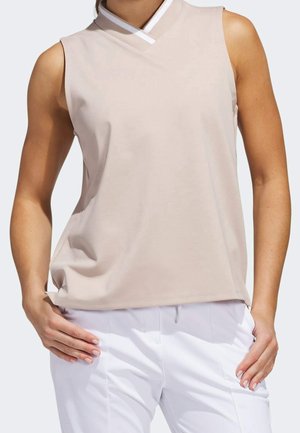 Ärmelloses beige Top mit V-Ausschnitt und weißem Besatz am Kragen, getragen zu weißen Hosen, Hände auf Hüfthöhe auf die Hüften gestützt.