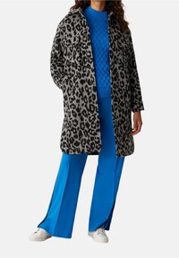Leopardmönstrad kappa i grått och svart, med två framfickor, som bärs över en blå kabelstickad tröja och blå vida byxor.