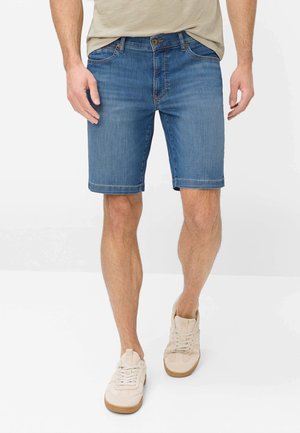 Mann trägt blaue Jeansshorts, beige T-Shirt und beige Sneakers, steht vor weißem Hintergrund.