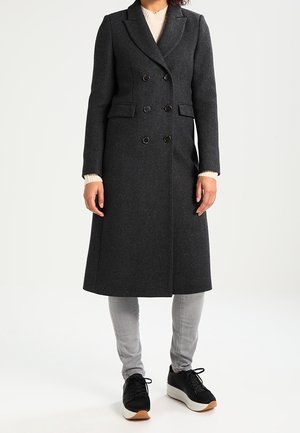 Femme portant un long manteau croisé gris foncé, un pantalon gris clair, des baskets noires et un pull beige en dessous.