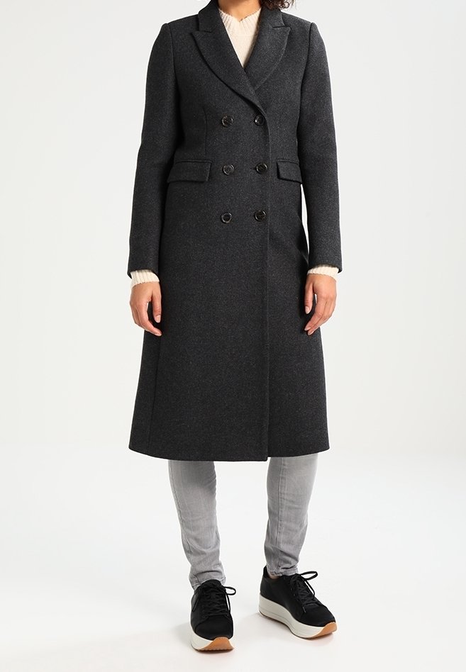 Femme portant un long manteau croisé gris foncé, un pantalon gris clair, des baskets noires et un pull beige en dessous.
