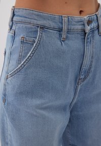 Jeans de mezclilla en azul claro con cintura alta, diseño de cinco bolsillos y detalle de costura en naranja contrastante. La tela parece suave con una ligera textura.