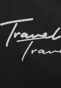 Černá látka s texturovaným bílým logem, "Travel" psaným kurzívou, ozdobeným malými stříbrnými tečkami pro detail.