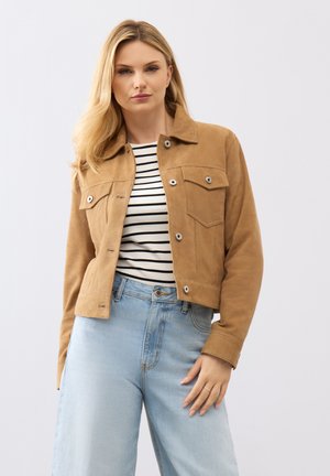 Blonde vrouw draagt een beige jas met knopen over een zwart-wit gestreept shirt, gecombineerd met lichtblauwe, high-waisted spijkerbroek tegen een effen achtergrond.
