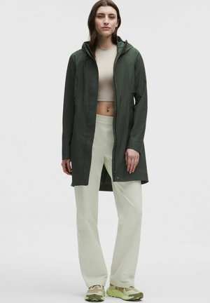 Mujer con chaqueta verde oscuro con capucha hasta la rodilla, top corto beige, pantalones anchos beige claros y zapatillas beige, de pie sobre fondo blanco.