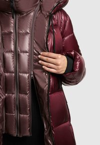 Personne portant et en train de dézipper partiellement une doudoune brillante bordeaux avec une couche intérieure zippée séparée en dessous.