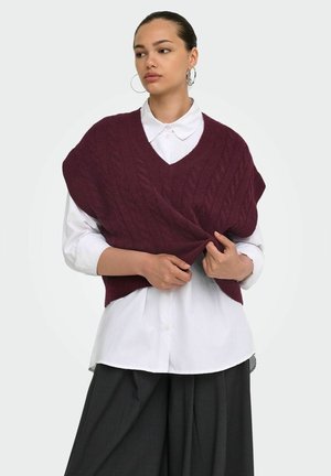 Femme portant une chemise blanche boutonnée sous un gilet sans manches bordeaux en maille torsadée, tenant l'ourlet du gilet avec les deux mains.
