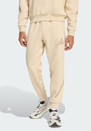 Uomo con pantaloni della tuta beige Adidas e felpa abbinata con scarpe sportive bianche e nere, in piedi su uno sfondo chiaro.