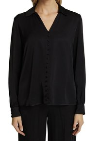 Femme portant un chemisier noir à manches longues avec des boutons recouverts de tissu sur le devant et un col, associé à un pantalon noir.