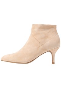 Botas de tobillo de ante beige con punta afilada y un tacón modesto. Textura suave con un diseño asimétrico y detalles de costura mínimos.