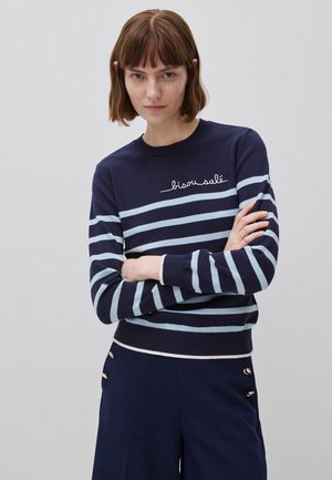 Mujer con cabello castaño corto que lleva un suéter azul marino con rayas horizontales azul claro y texto blanco que dice "bisou salé", combinado con pantalones azul marino con botones dorados.