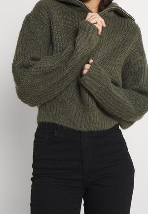 Person iført en chunky olivengrøn lynlåstrøje med oversize ærmer og sorte højtaljede bukser, hænderne nær trøjes krave.
