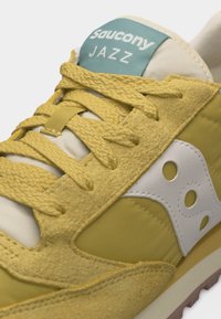 Κοντινό πλάνο κίτρινου αθλητικού παπουτσιού Saucony Jazz με υφασμένες κορδόνια, σουέτ επικαλύψεις και λευκό πάνελ με λογότυπο στο πλάι.