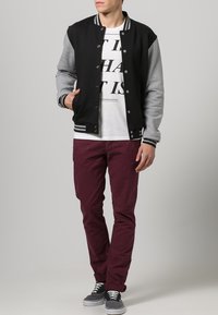 Zwart en grijs varsityjack, grijze mouwen, gestreepte kraag, gecombineerd met een wit shirt, bordeauxkleurige broek en grijze sneakers.