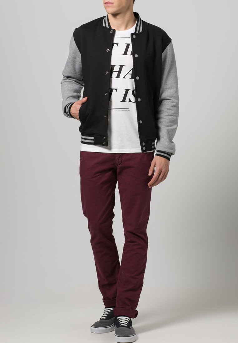 Zwart en grijs varsityjack, grijze mouwen, gestreepte kraag, gecombineerd met een wit shirt, bordeauxkleurige broek en grijze sneakers.